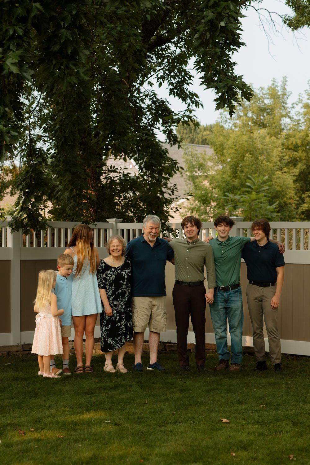 A family stands together on a green lawn in front of a white fence during a warm summer evening.