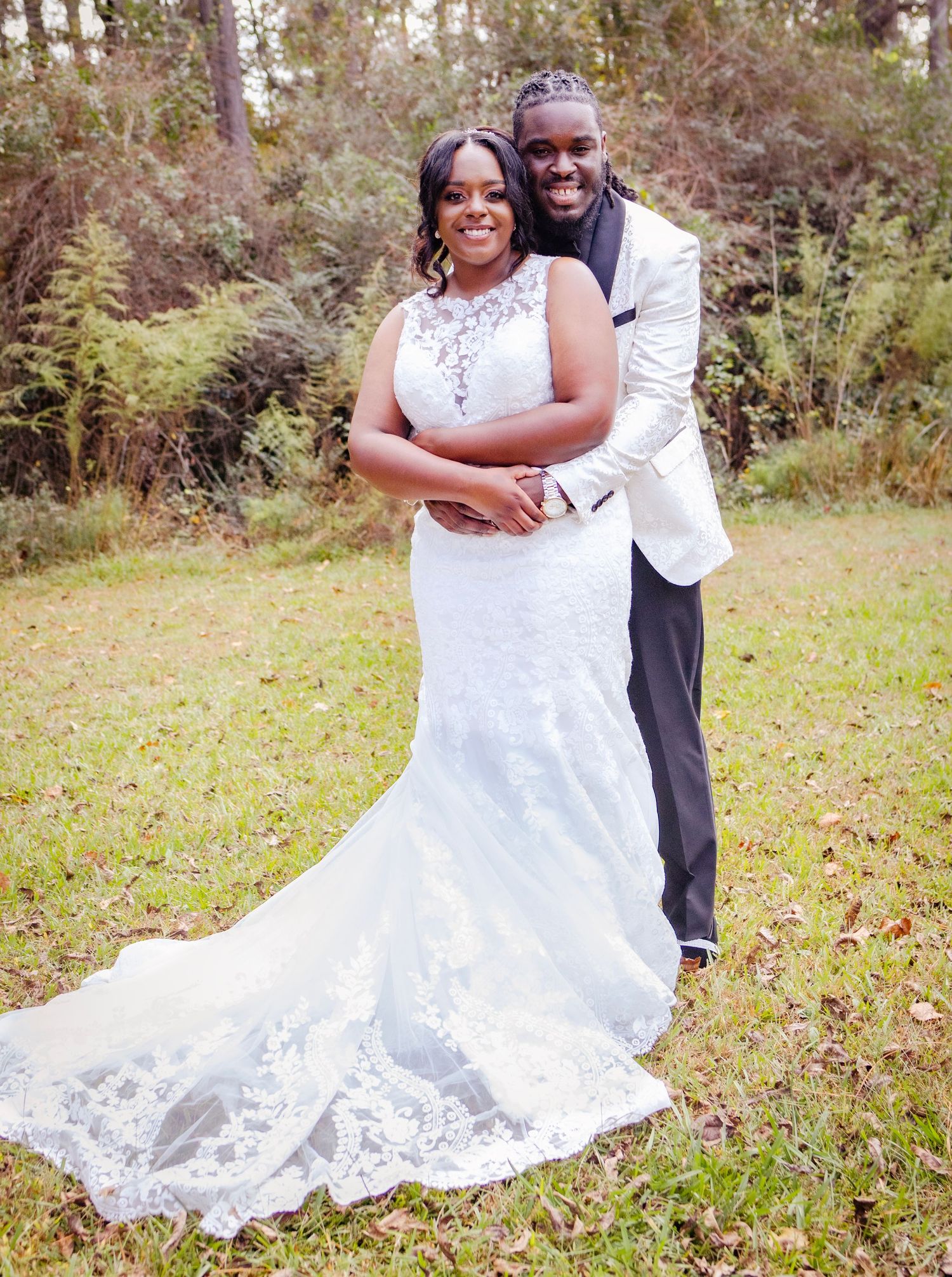 A couple in wedding attire shares a joyful embrace while standing in a grassy autumn meadow at sunset.