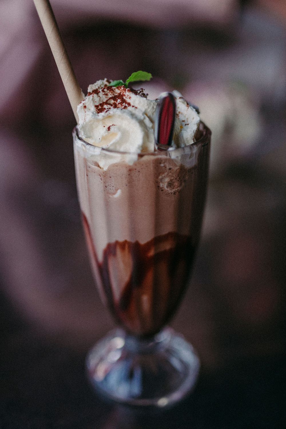 A chocolate milkshake topped with whipped cream, chocolate sprinkles and a red straw in a tall sundae glass.