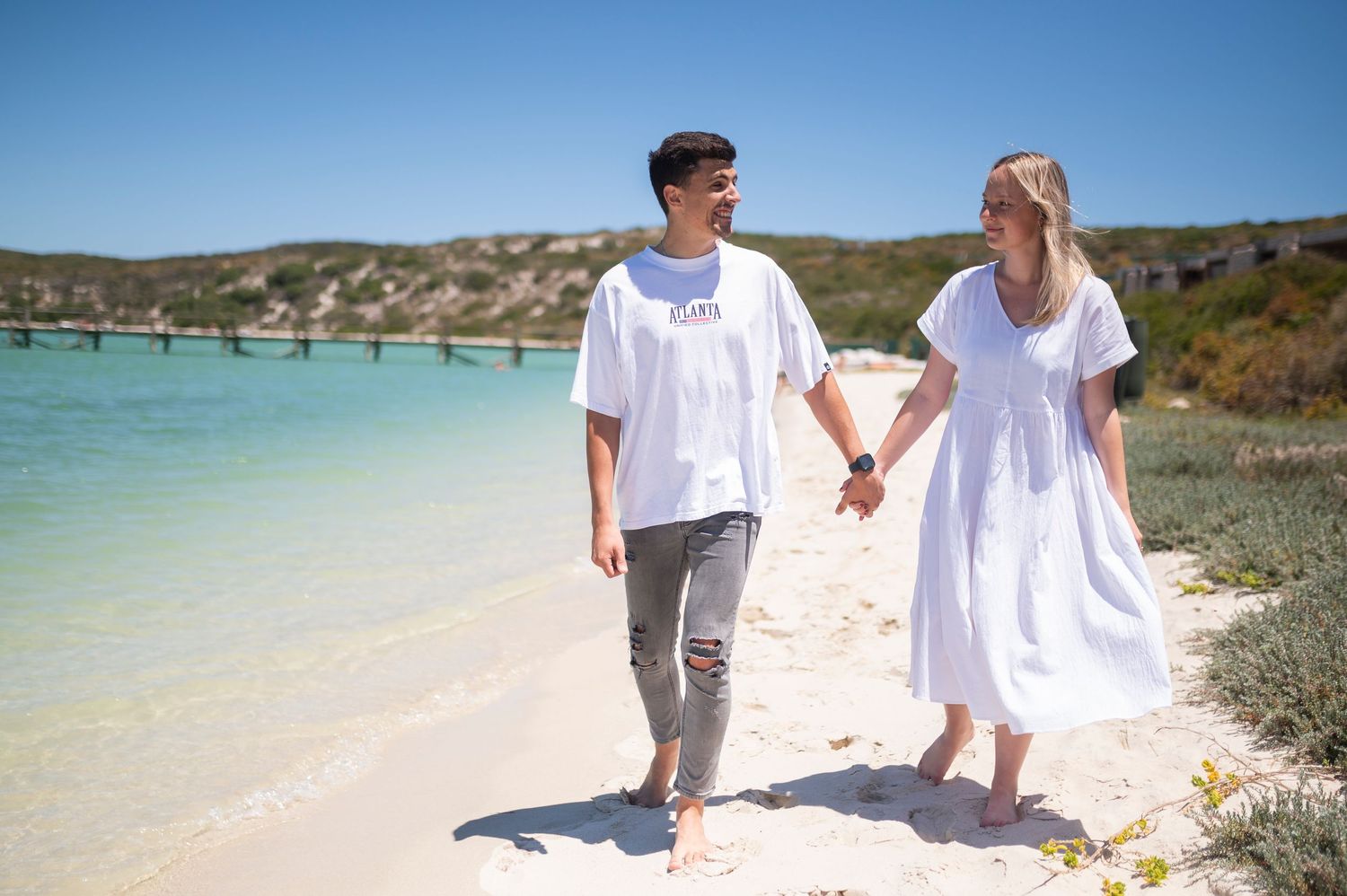 Couple in white outfits walking and holding hands along a scenic beach shoreline during sunset.