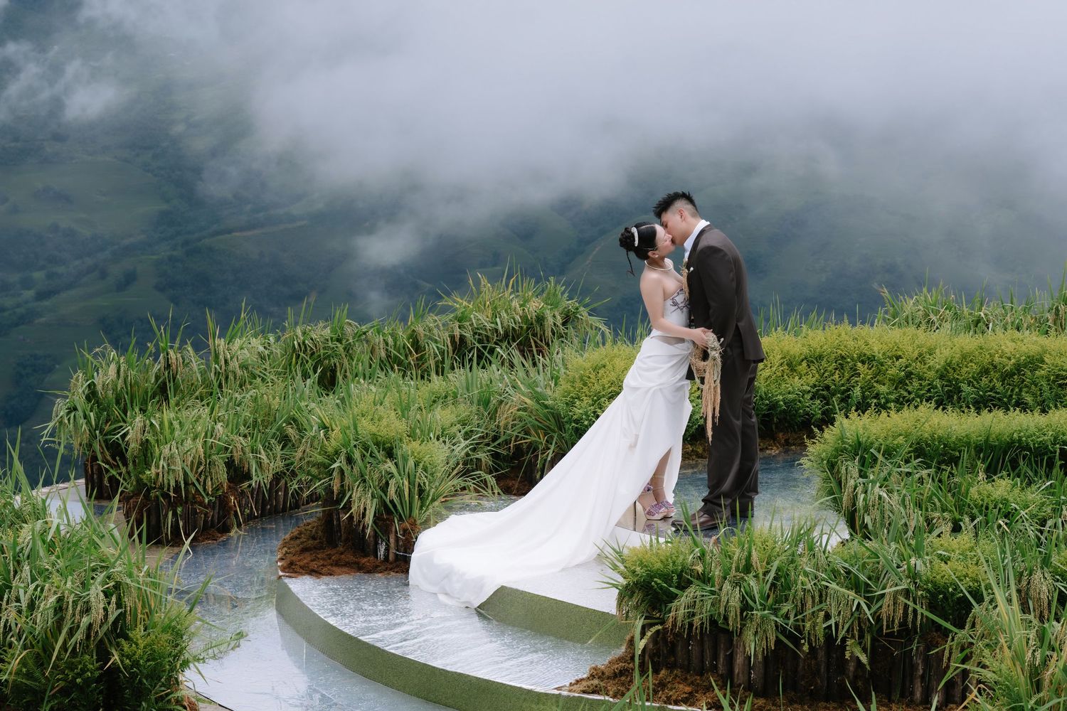 Bride and groom sharing a romantic kiss on a misty mountain terrace surrounded by lush green rice terraces.