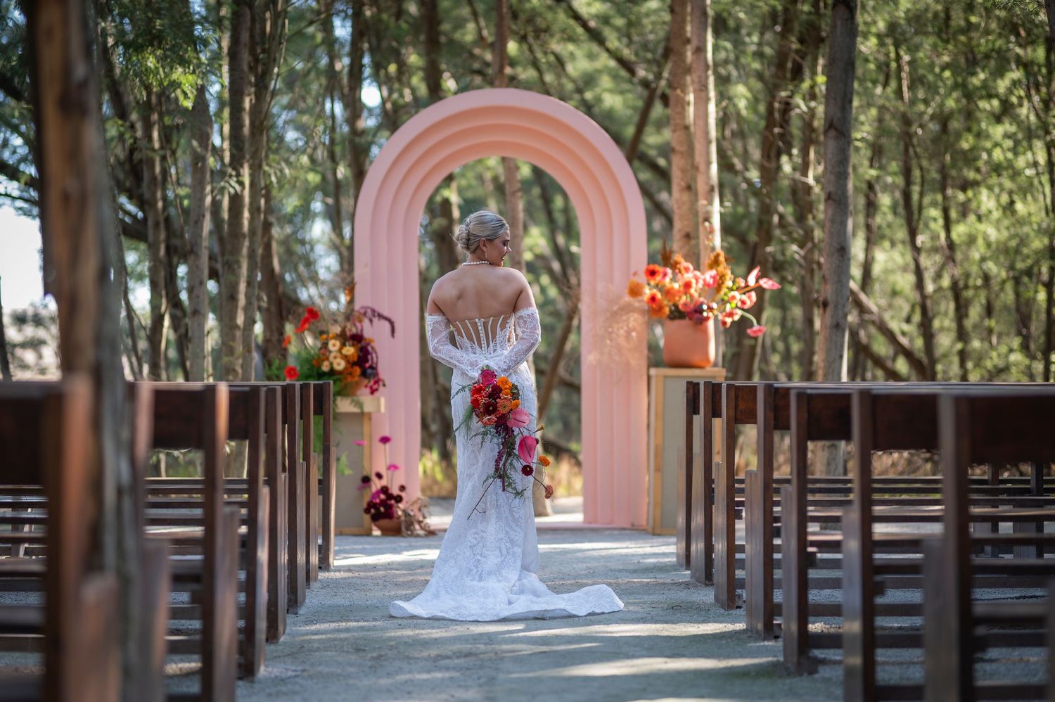 Outdoor wedding ceremony setup in forest with pink archway and wooden pews decorated with autumn floral arrangements.