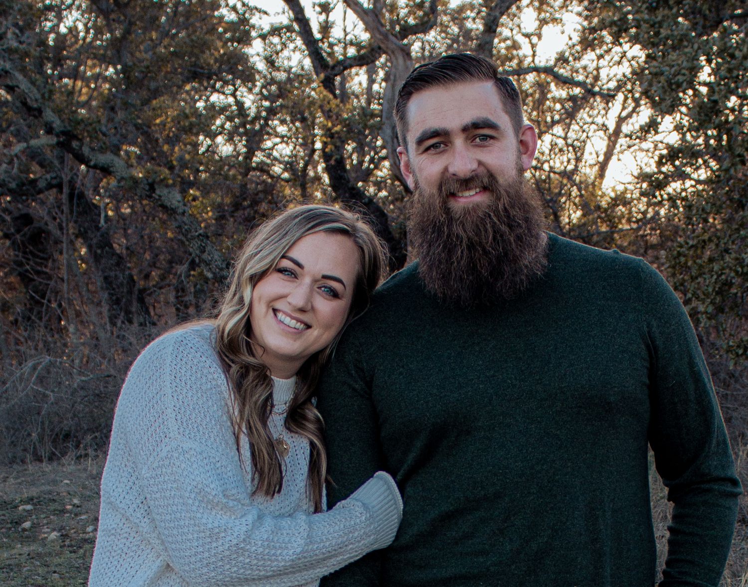 A couple smiles together outdoors at sunset, with autumn foliage in the background.