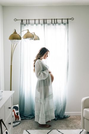 A bride in an ethereal white wedding dress and veil stands in a brightly lit room with muted teal curtains and warm lighting.