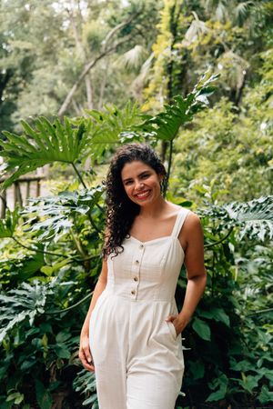 Person in a white sleeveless dress poses naturally against lush green foliage background in a garden setting.