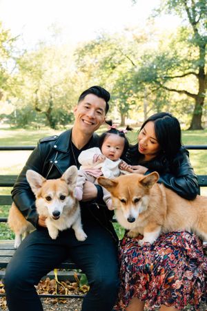 A family sits on a park bench with their two Corgi dogs on a sunny autumn day.