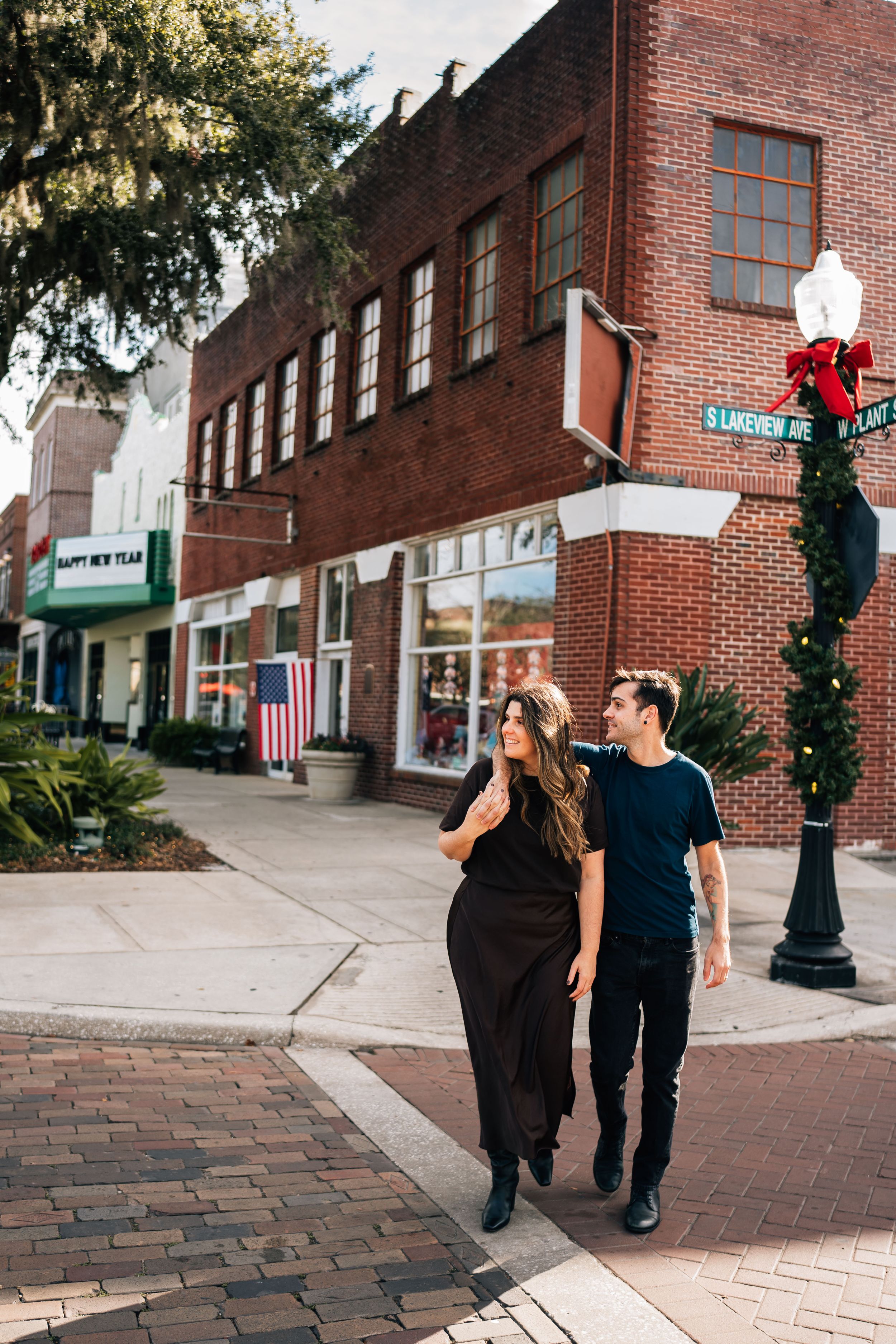Two people stroll along a charming brick sidewalk in a historic downtown area with red brick buildings and street lamps.
