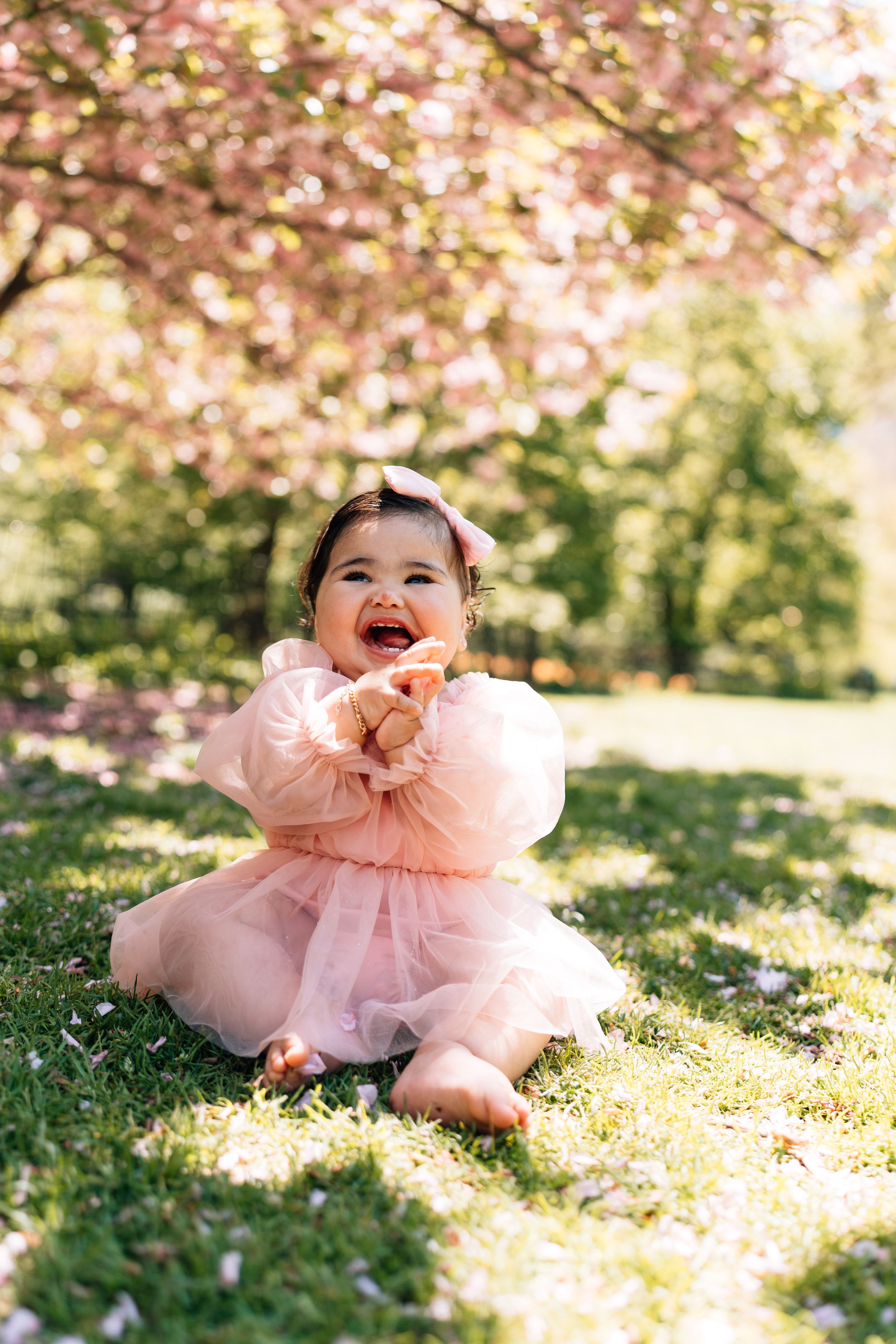 A delightful scene featuring a baby in pink tulle dress laying on grass beneath cherry blossoms.