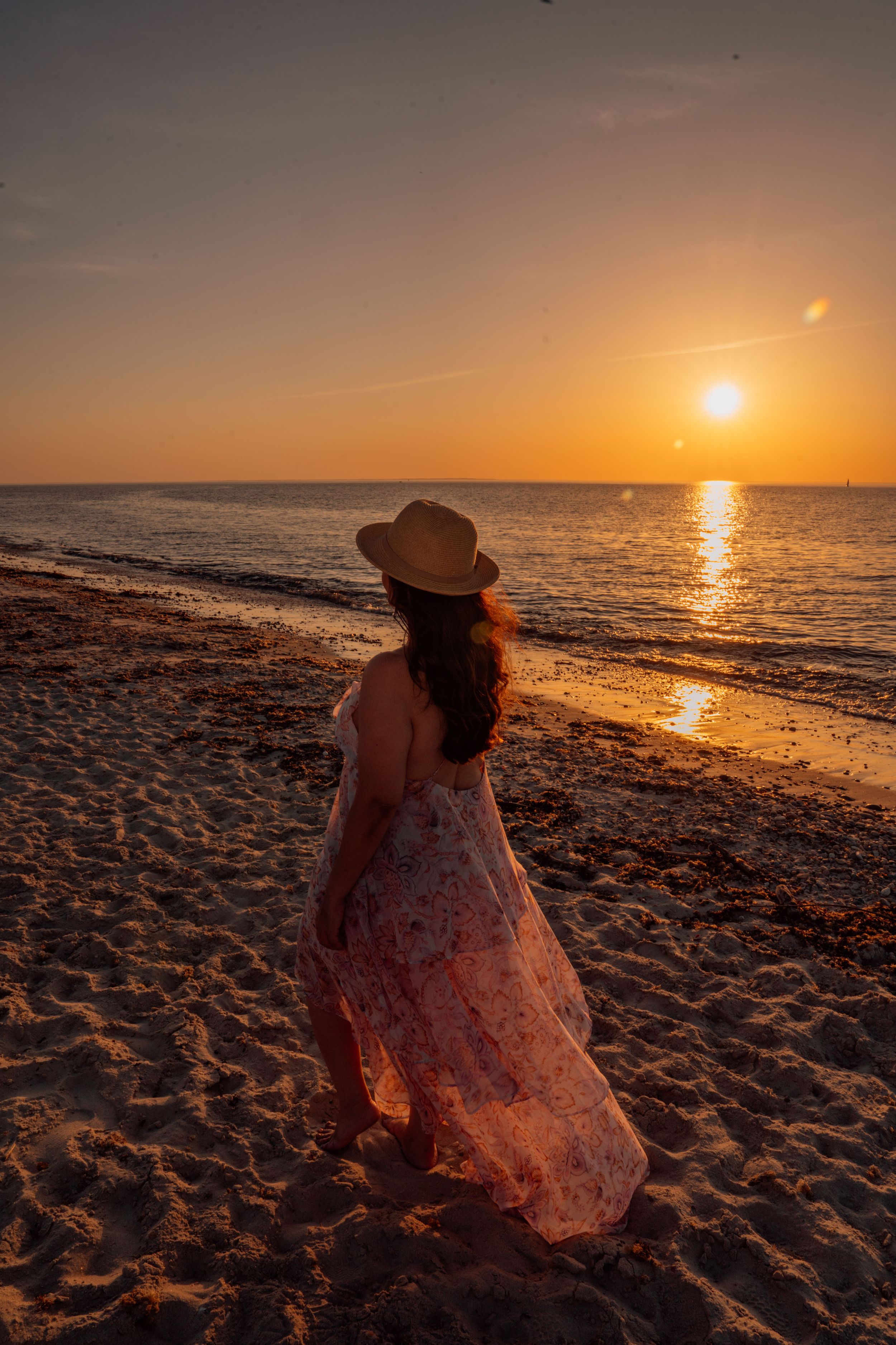 A figure in a flowing dress sits on a sandy beach watching the golden sunset over the ocean horizon.