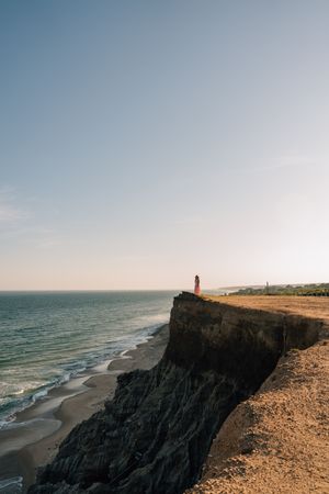 A dramatic rocky coastline with steep cliffs overlooking an ocean beach at sunset with a distant lighthouse on the horizon.