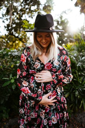Beautiful floral dress and wide-brim black hat shown in outdoor sunlit maternity photo shoot with garden backdrop.