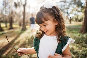 A little child explores nature outdoors in a sunny garden while wearing a white dress and floral accessories.