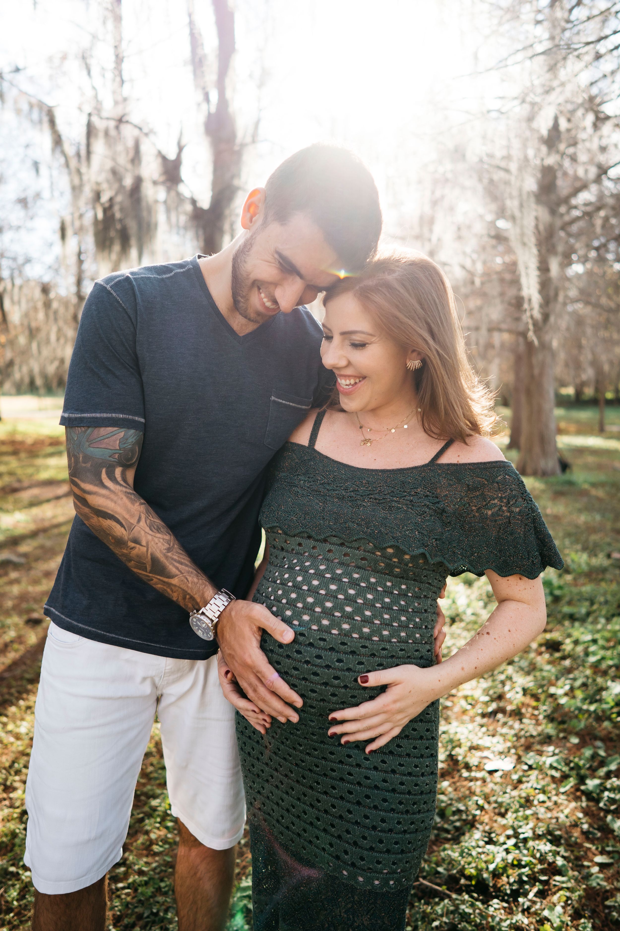 A couple embraces during a maternity photo session in a sunlit wooded area on a bright afternoon.