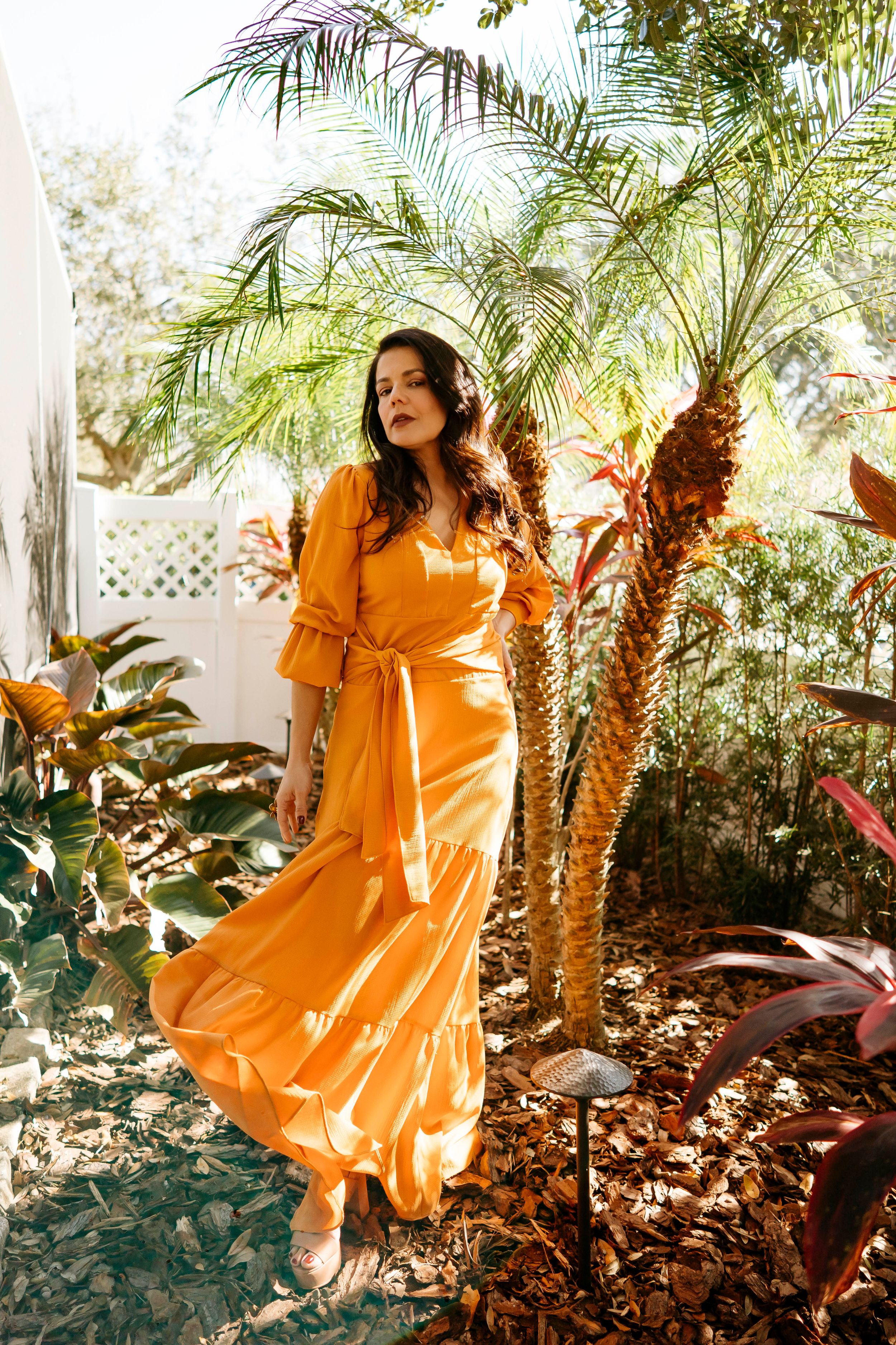 A woman in a flowing orange maxi dress dances and twirls in a lush tropical garden setting.