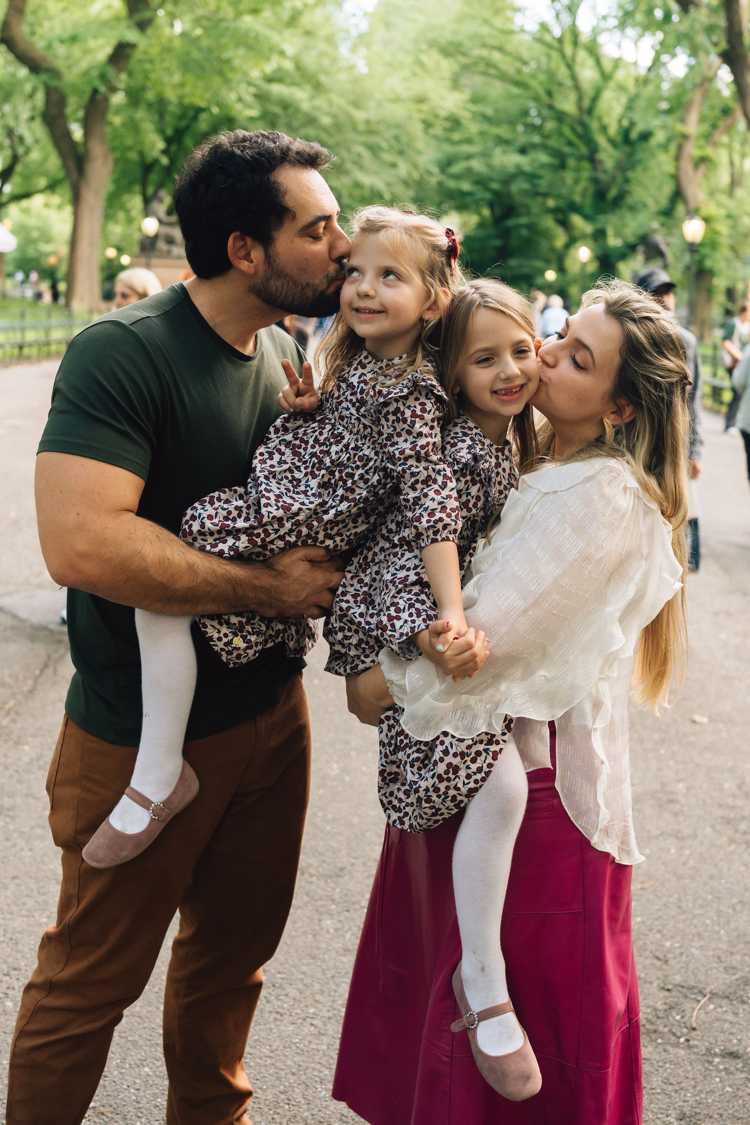 Family wearing coordinated outfits shares affectionate moment on tree-lined path in Central Park.