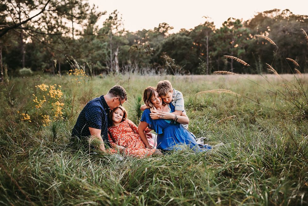 A loving family shares tender moments together while sitting in a sunlit field surrounded by tall grass and trees.