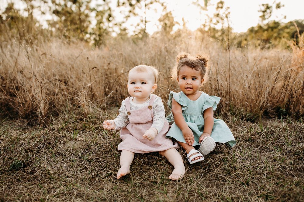 Two children in light pastel colored outfits sit together in a grassy meadow during golden hour sunset.