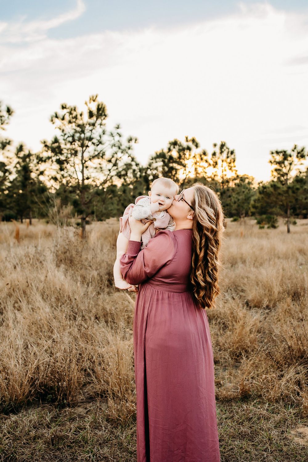 Rustic outdoor portrait session in a golden field with dried grass and pine trees at sunset.