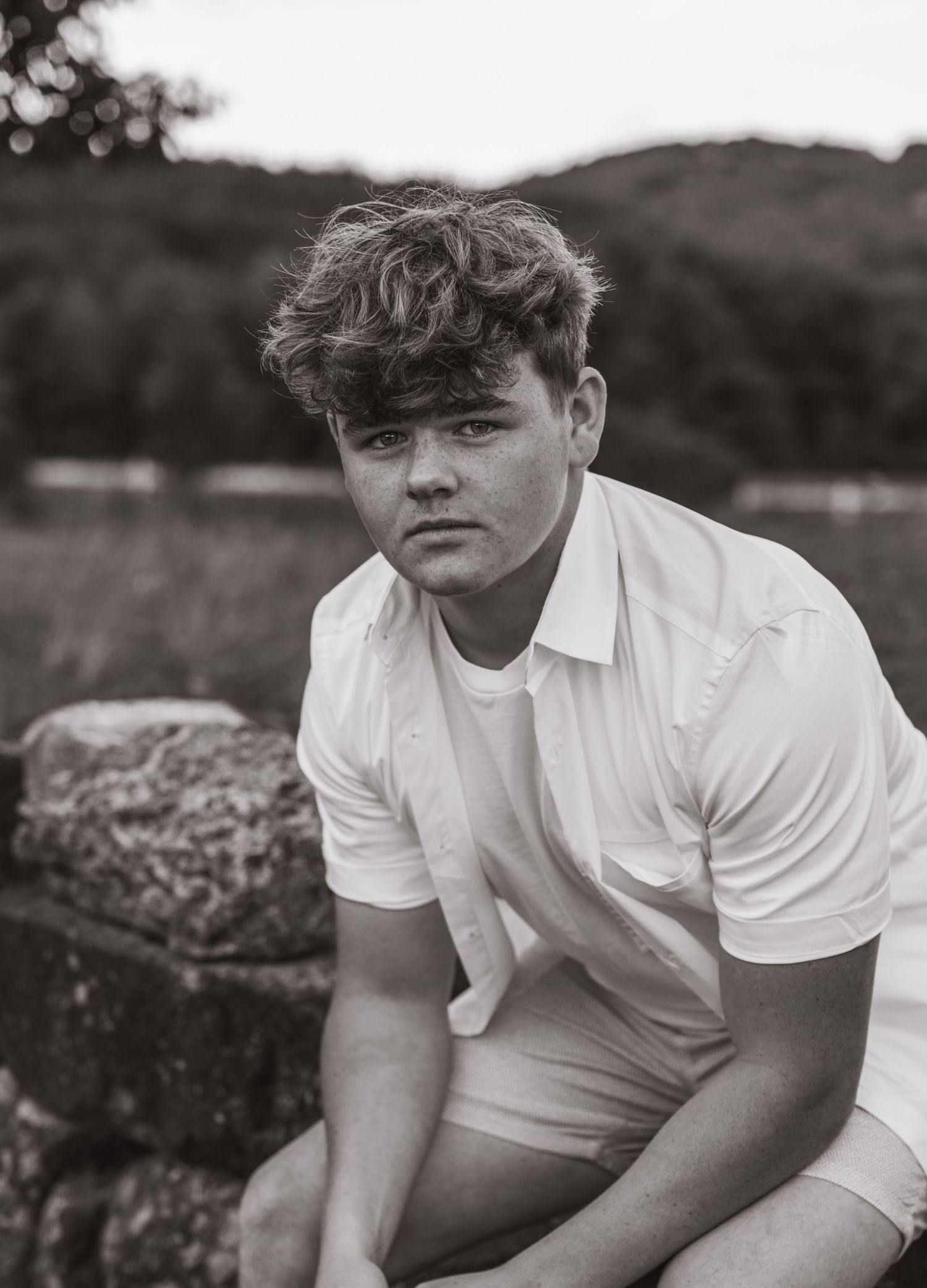 A young man in a white shirt sits on a stone wall in a black and white portrait.