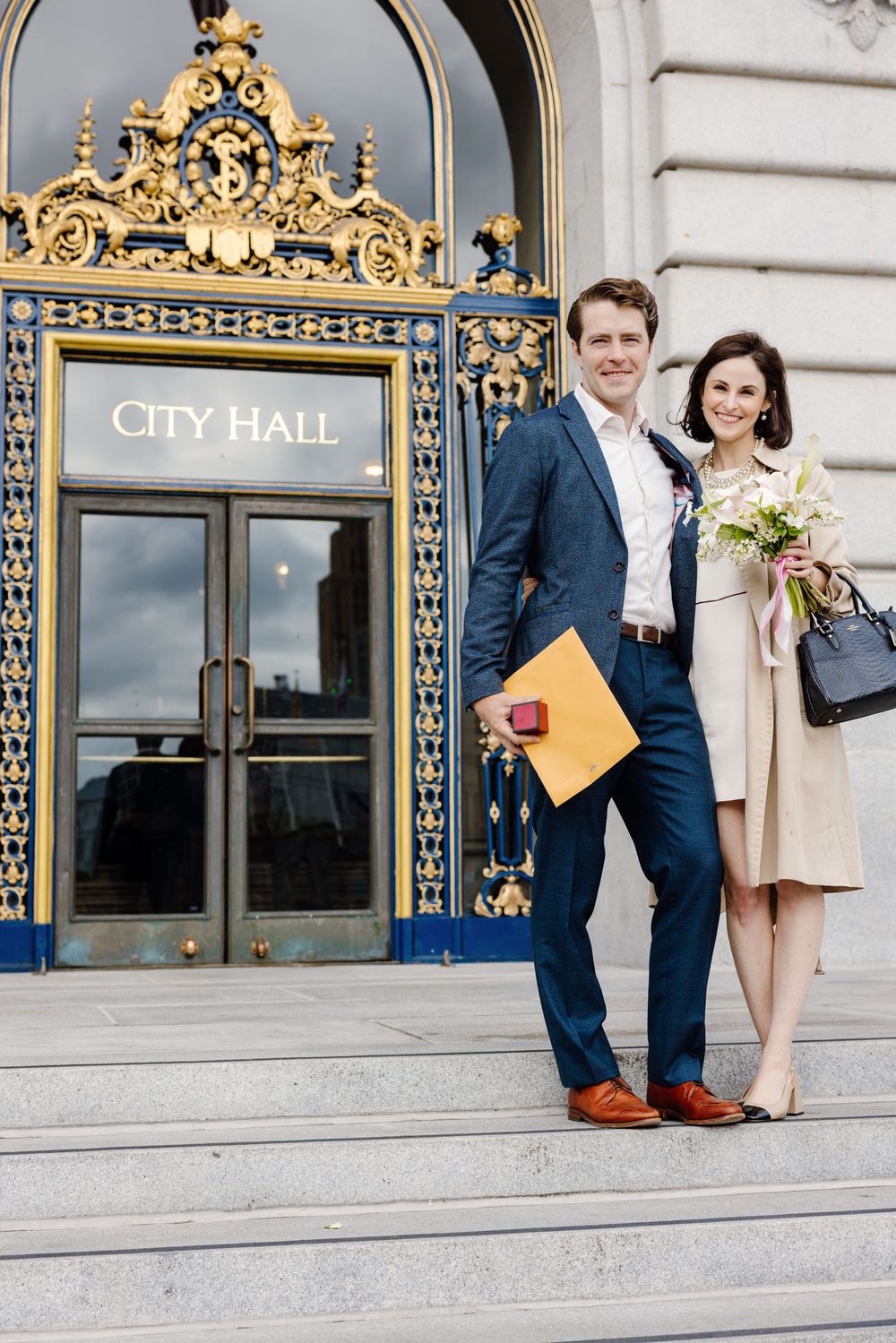 A couple stands together outside city hall on a sunny day with ornate architectural details in the background.
