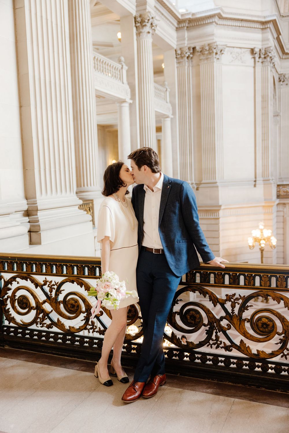 A romantic couple shares tender moments along an ornate marble balustrade in a grand historic building with classic columns.