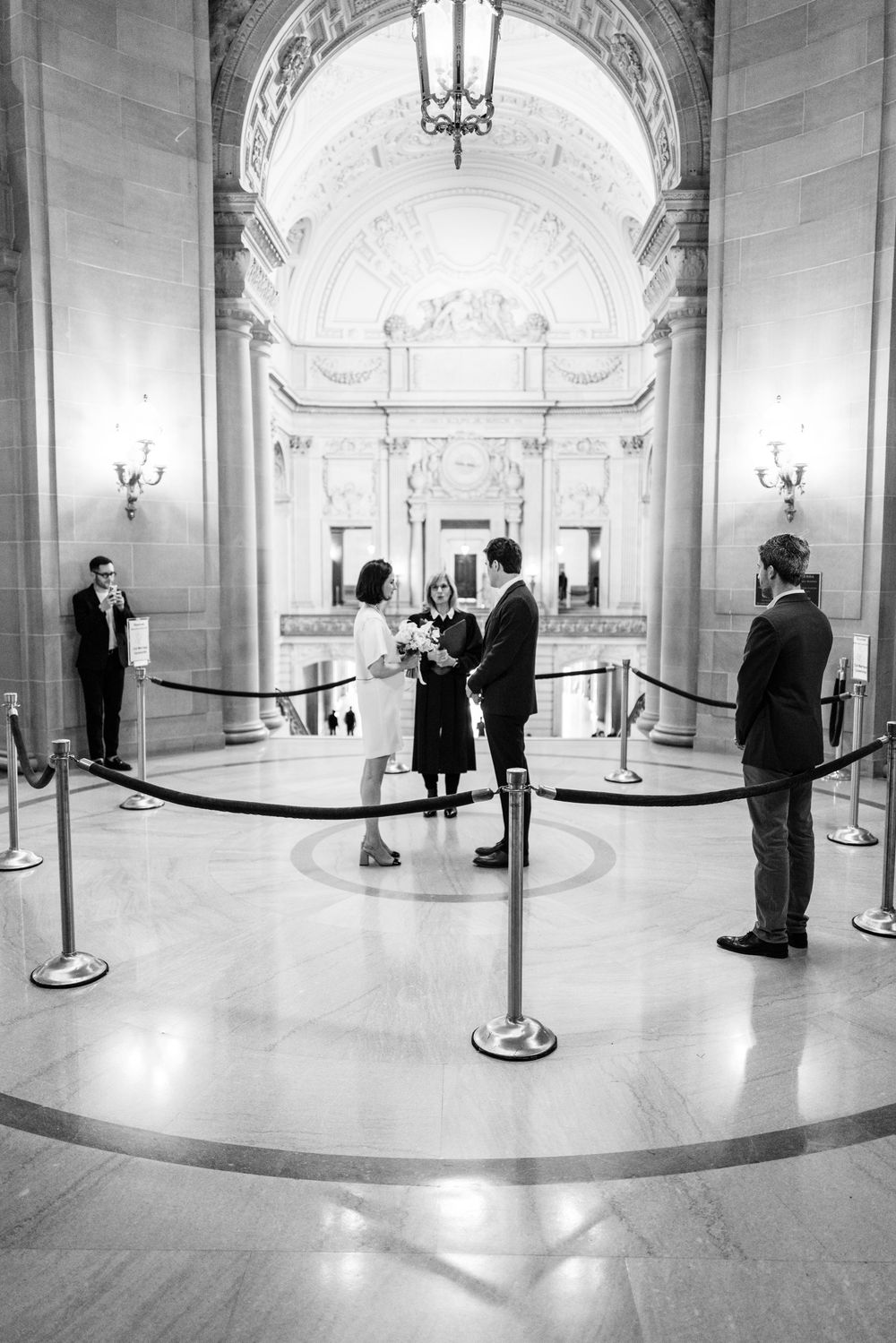 Black and white panoramic view of a grand marble hallway with stanchions and people waiting.