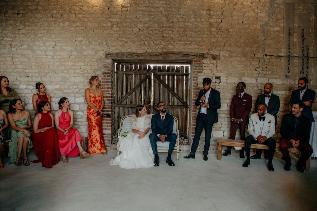 Bride and groom seated at ceremony surrounded by diverse wedding guests in a rustic barn.