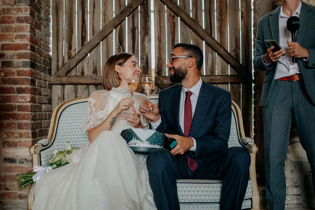 Bride and groom toast with champagne while seated on a vintage sofa during wedding reception.