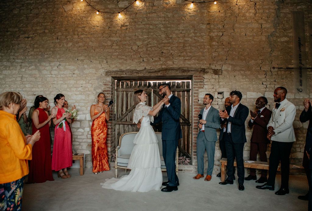 Bride and groom share first kiss surrounded by cheering guests in a rustic barn ceremony.