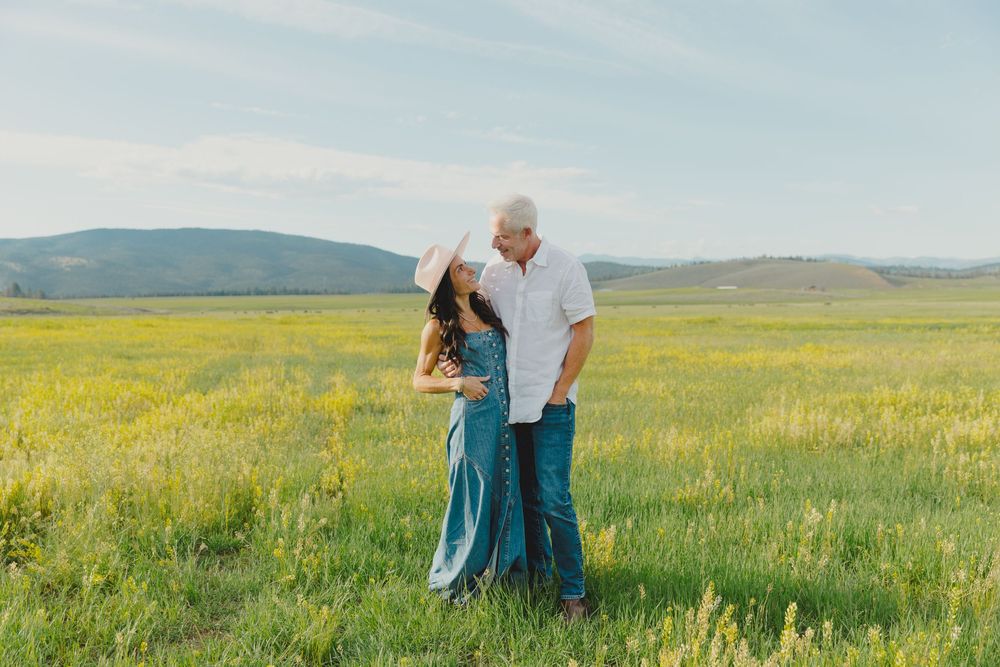 Couple standing in a lush green field with yellow wildflowers and rolling hills under a blue sky.
