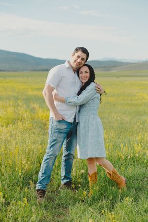 A couple embracing in a sunny green field with yellow wildflowers and mountains in the background.