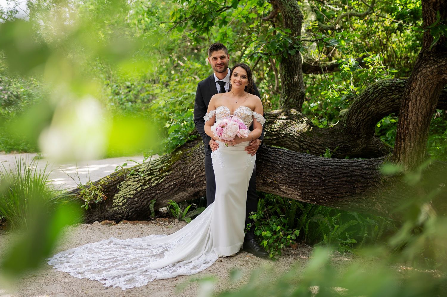 A romantic wedding couple embraces in a lush garden setting with soft natural light filtering through the trees.