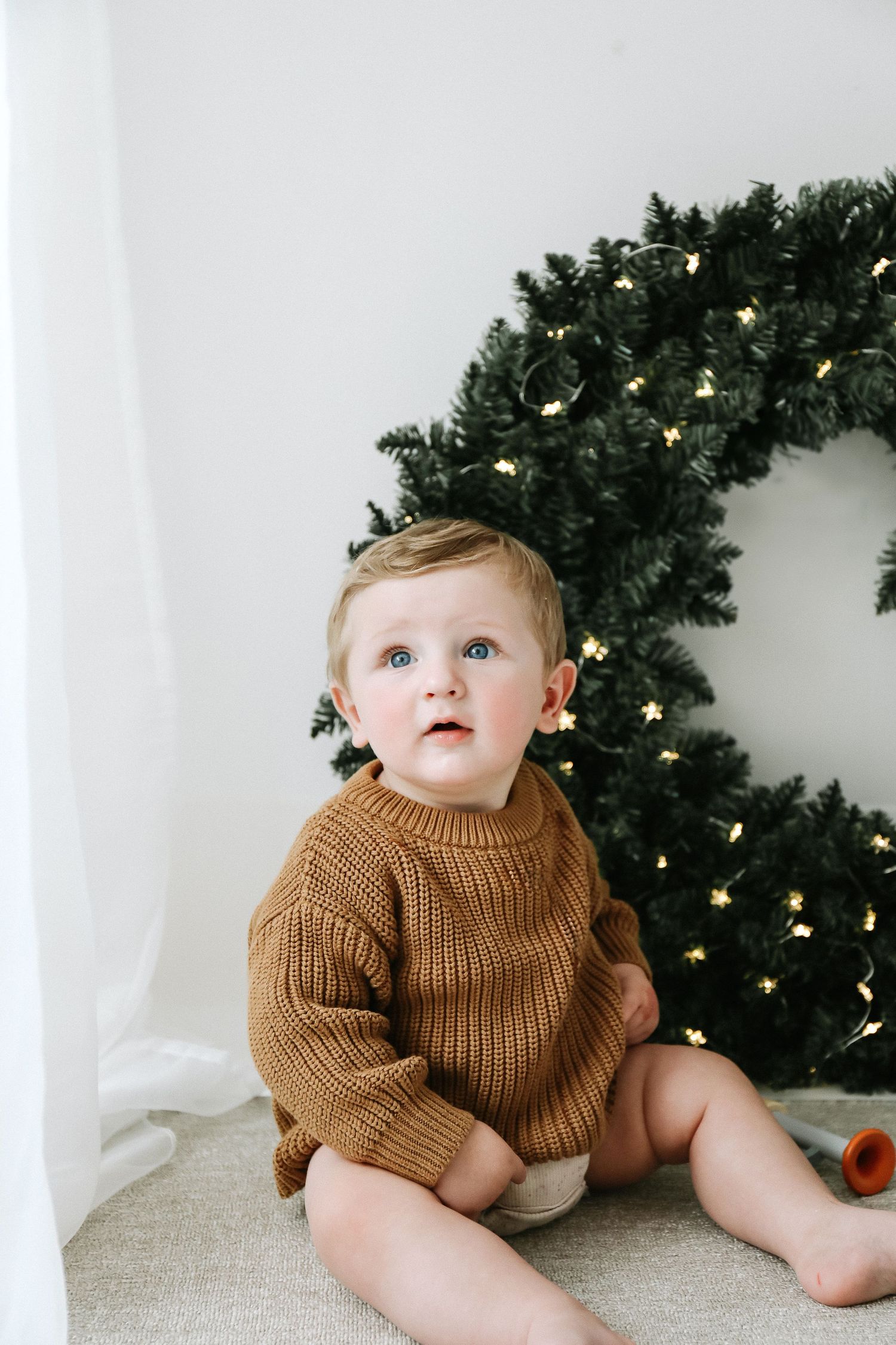 Baby wearing brown knit sweater sits in front of  Christmas wreath, laughing joyfully with twinkling lights during photoshoot in Nottingham