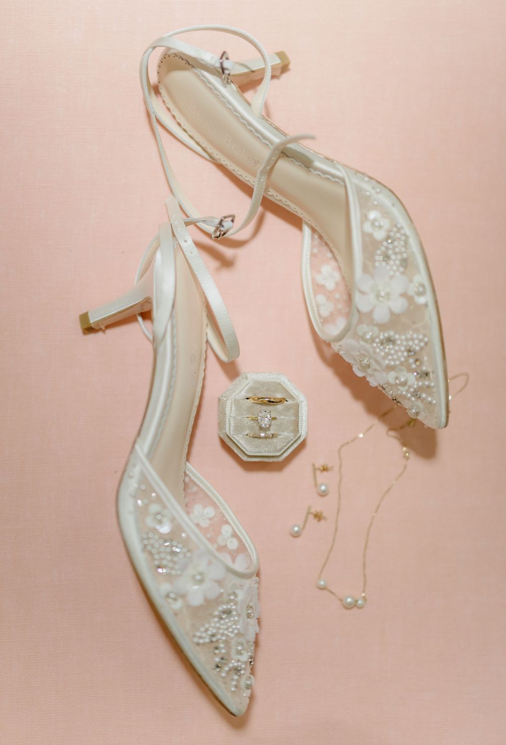 Elegant white wedding shoes with pearl and crystal embellishments displayed against a soft pink background.