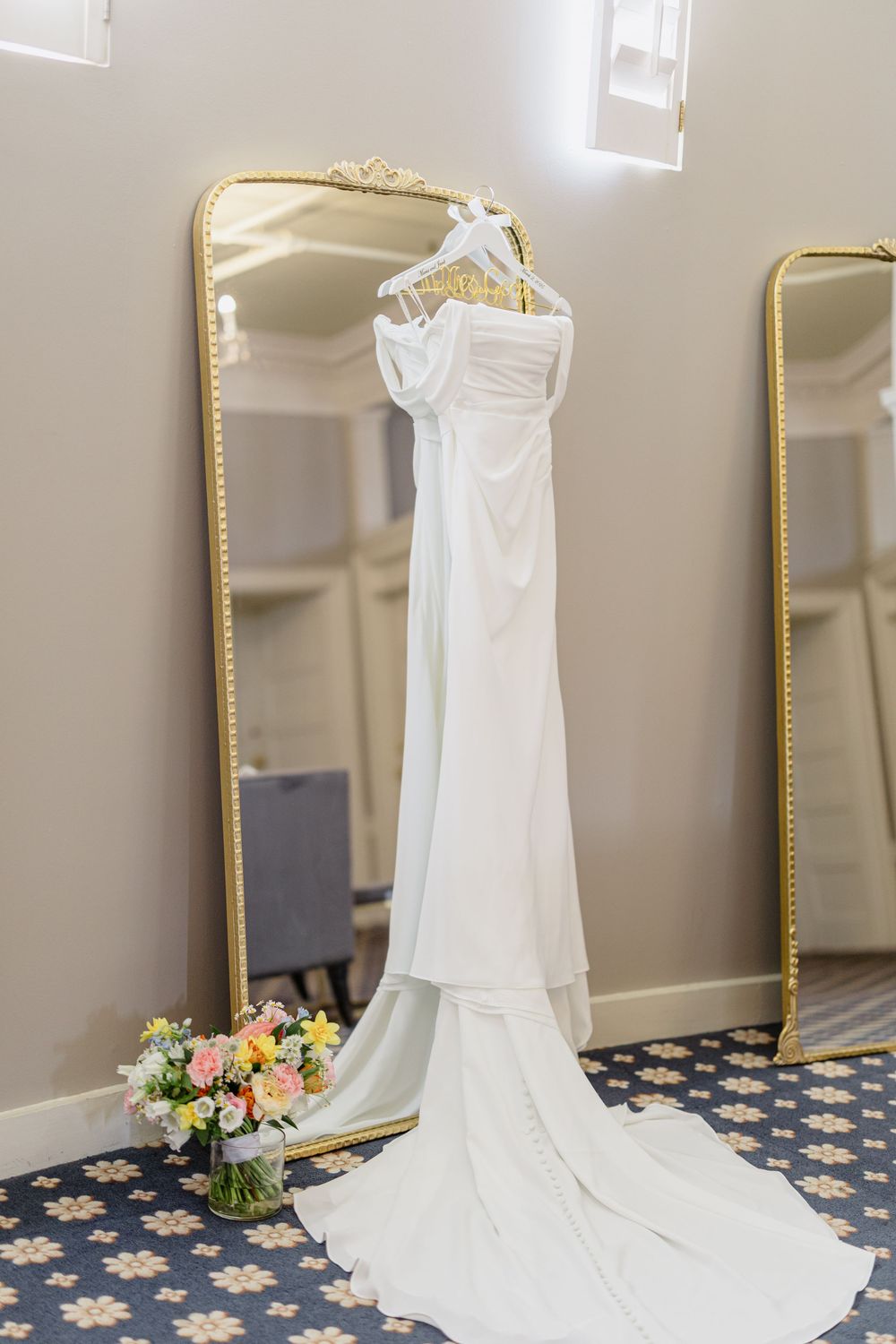 White wedding dress hanging on display with a long train against a neutral wall with gold-framed mirror.