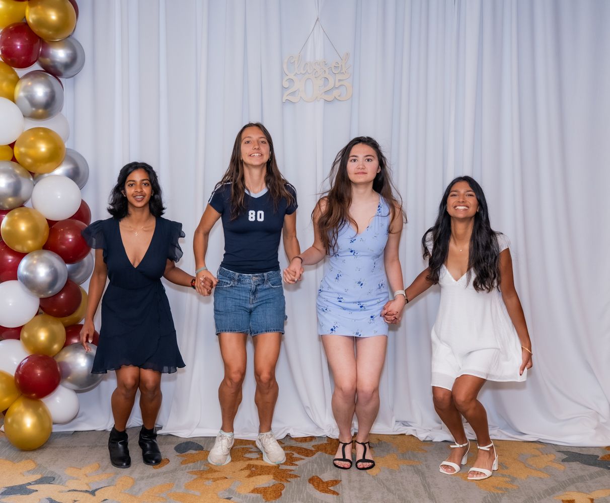 Four friends pose in casual attire against a white curtain backdrop with gold and burgundy balloons.