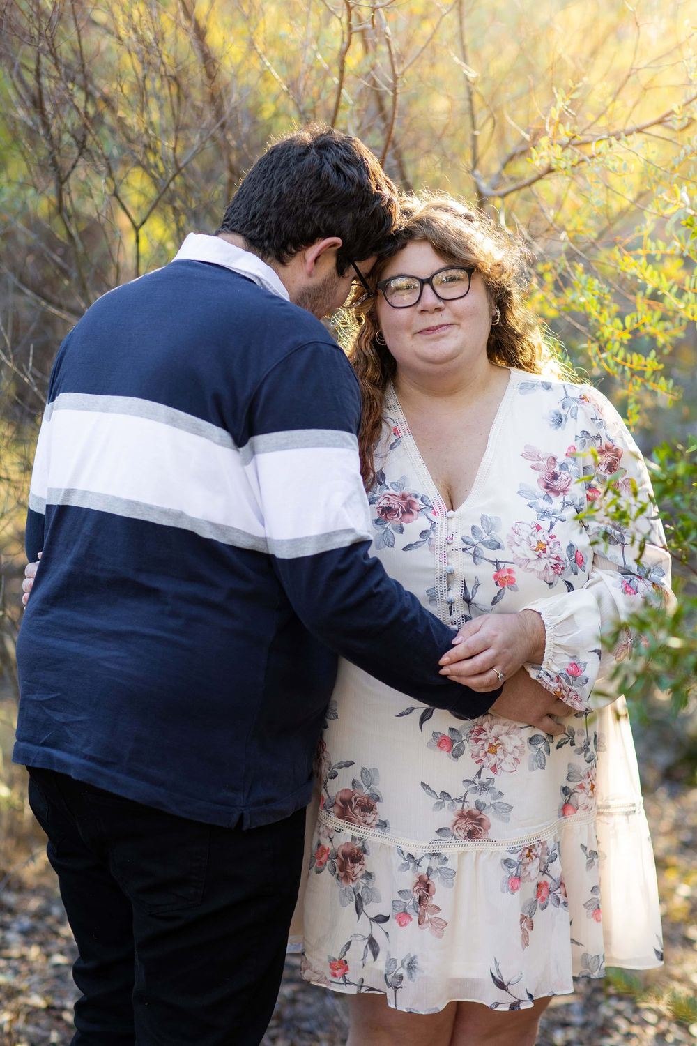 A couple embraces outdoors in autumn, wearing a navy striped sweater and floral dress against a golden-lit natural backdrop.