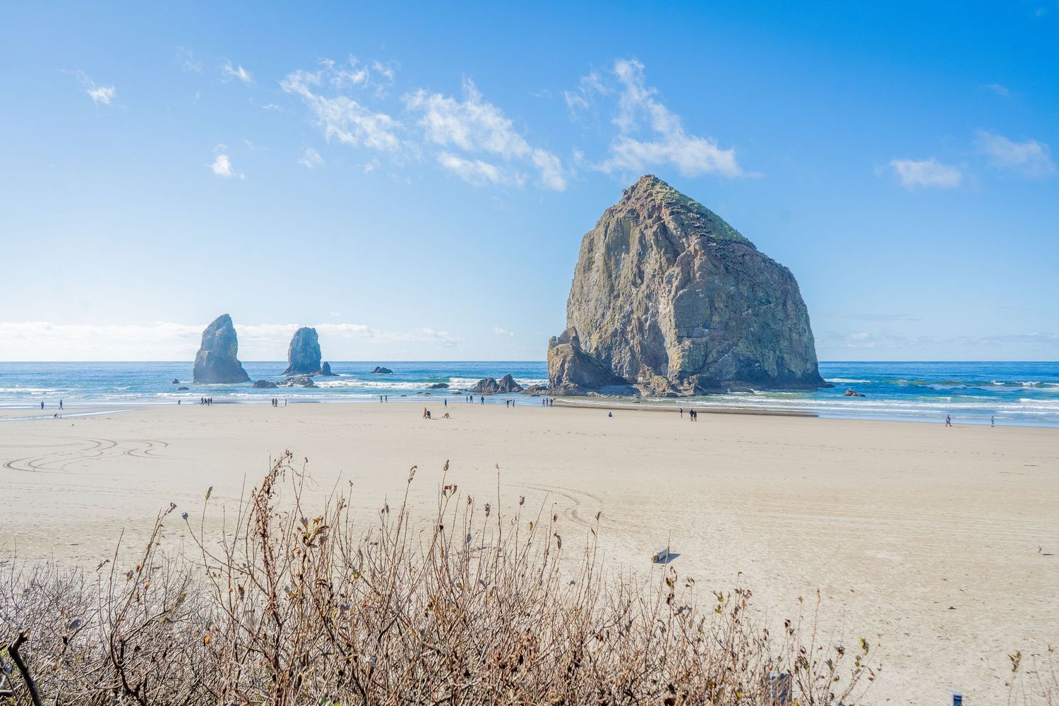 Scenic view of Haystack Rock and coastal rock formations along Cannon Beach under bright blue Oregon skies.