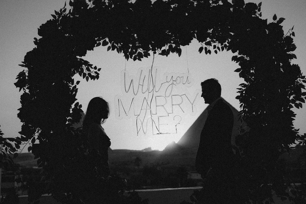 Black and white silhouette photograph showing two figures beneath an archway with illuminated text asking Will You Marry Me.