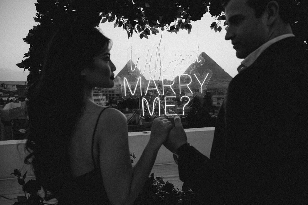 A romantic proposal scene captured in black and white silhouette against a dramatic sky background.