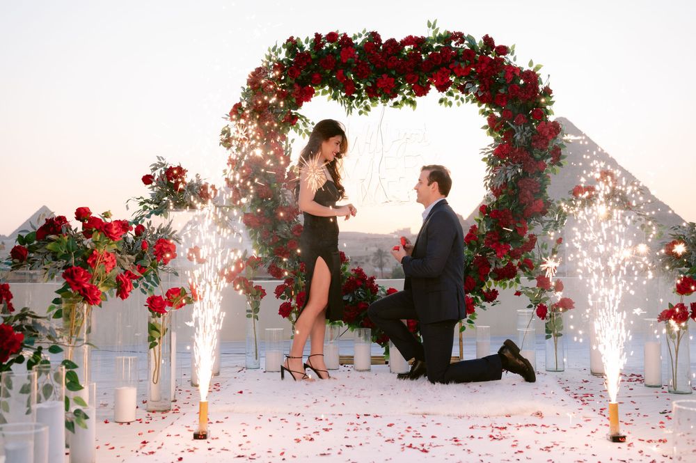 A proposal scene under a floral arch decorated with red roses and sparkling firework fountains.