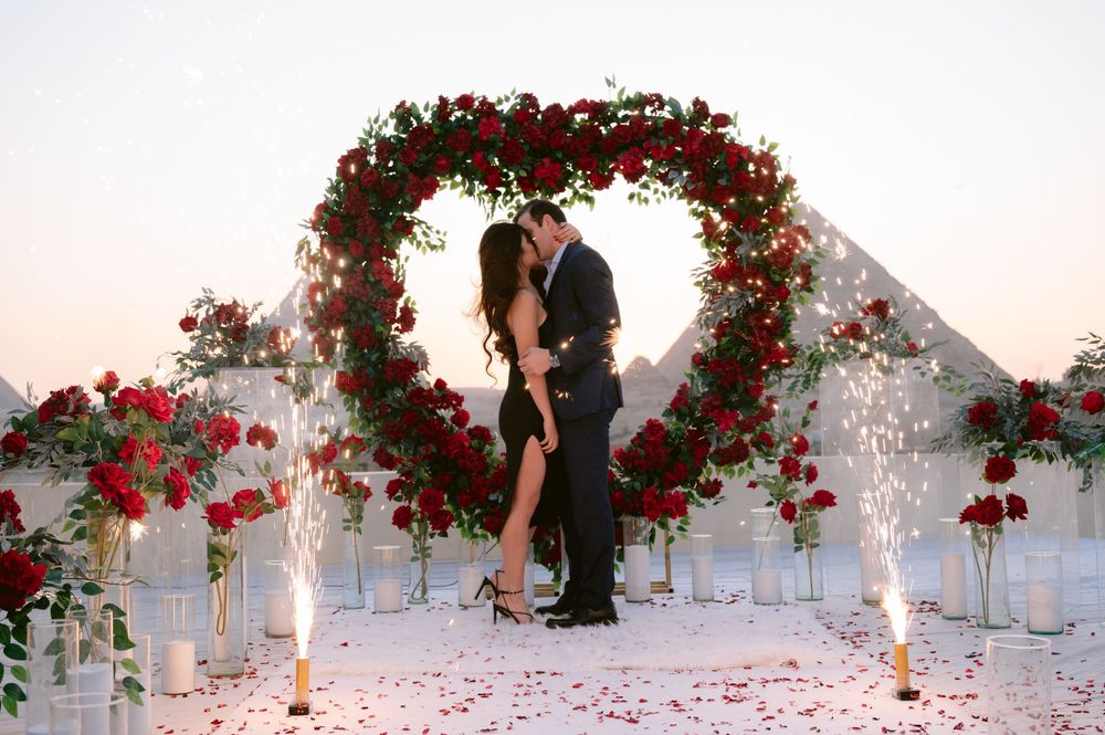 Romantic outdoor archway decorated with red roses and candles creating a dreamy wedding ceremony scene at sunset.