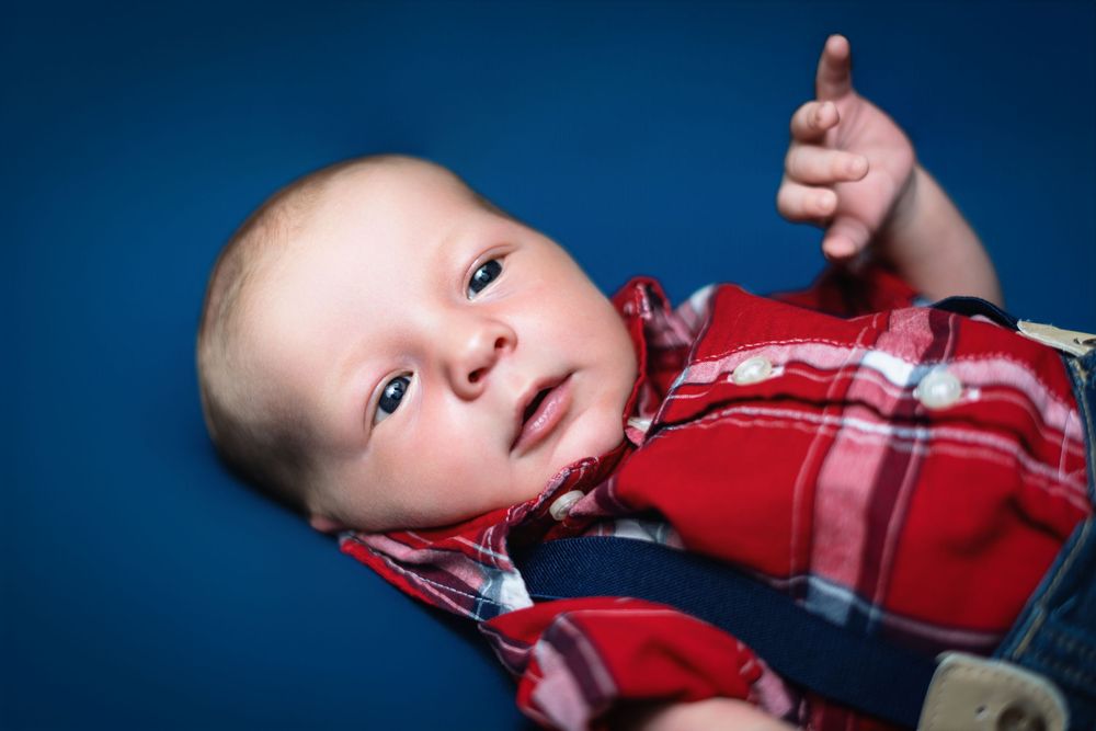Baby wearing red plaid shirt and denim overalls lying on blue background.