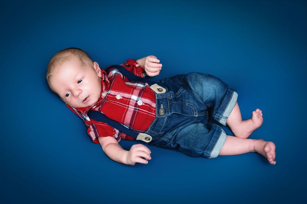 Infant in red and white plaid outfit lying on dark blue backdrop.