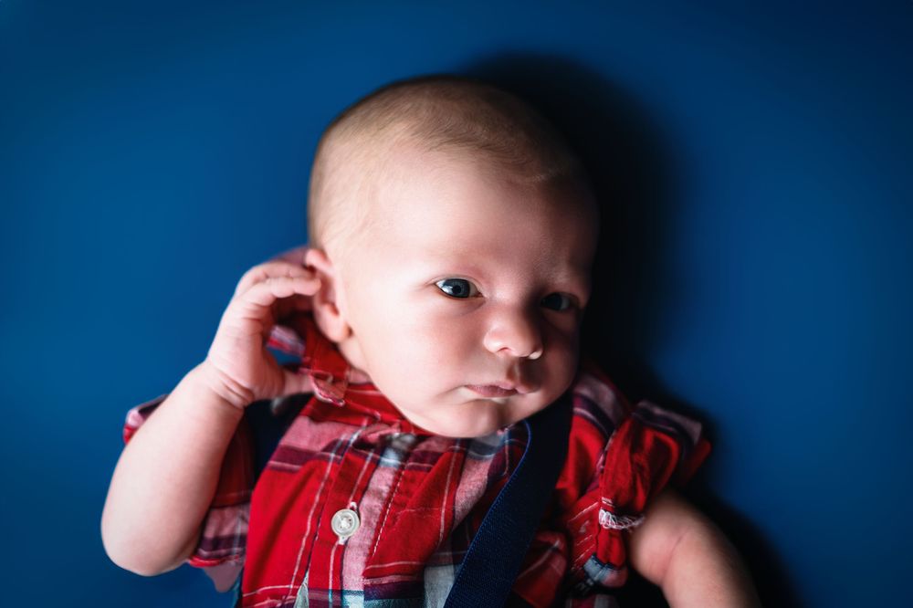 Baby in red plaid shirt and overalls resting on blue studio background.