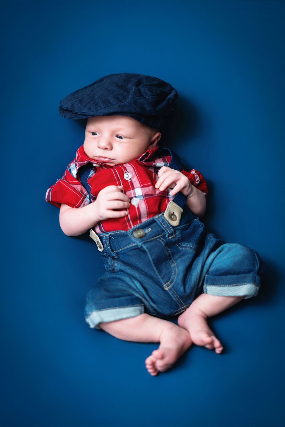 Baby wearing baseball cap, plaid shirt and denim overalls on blue background.