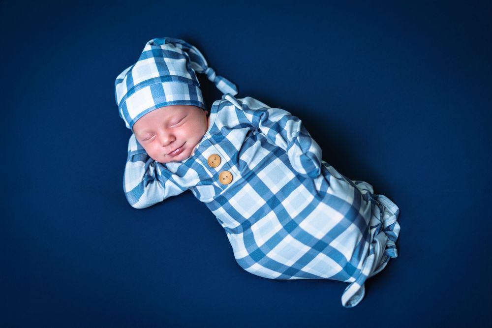 Newborn wearing blue and white checkered outfit sleeping against dark blue background.