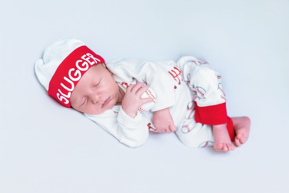 Newborn in white outfit and red baseball cap lying on white background.