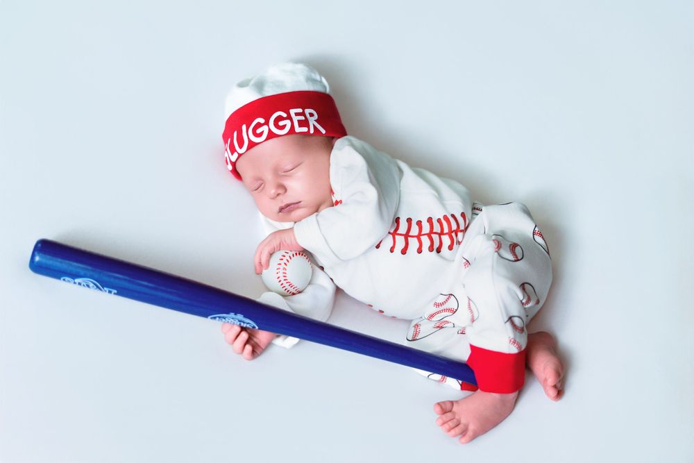Baby wearing baseball themed outfit with red cap and blue bat prop on white surface.