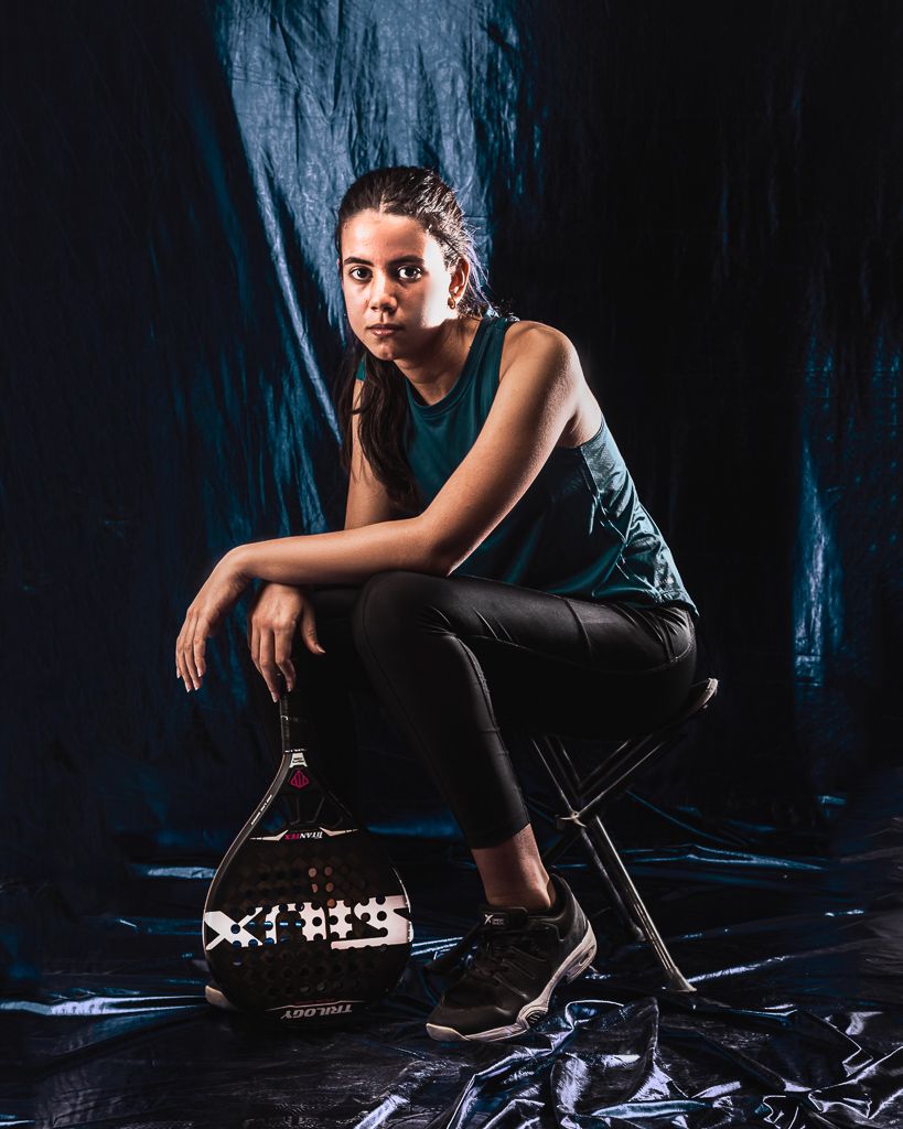 Athletic figure in dark clothing crouching with padel tennis racket in dramatic lighting.